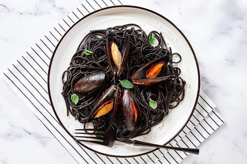 Black spaghetti with mussels, on a white plate with a fork, on a striped napkin, marble background, homemade, no people