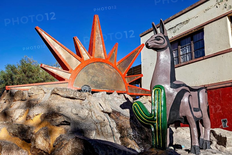Close-up view of a llama statue next to a sun symbol and cacti on a rocky ground, monument and statue in the streets of Calama in Chile