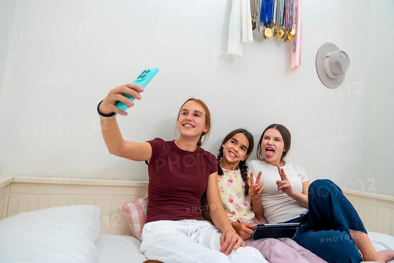 Teenage girl holding smartphone, capturing a moment with her two younger sisters making funny faces and peace signs while sitting casually on a bed in their bright bedroom