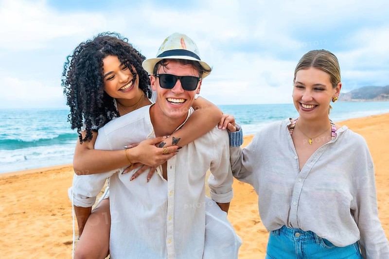 Diverse group of young adults laughing and hugging on a sunny beach, enjoying a carefree summer vacation together with playful piggyback rides, waves, sand and bright blue sky