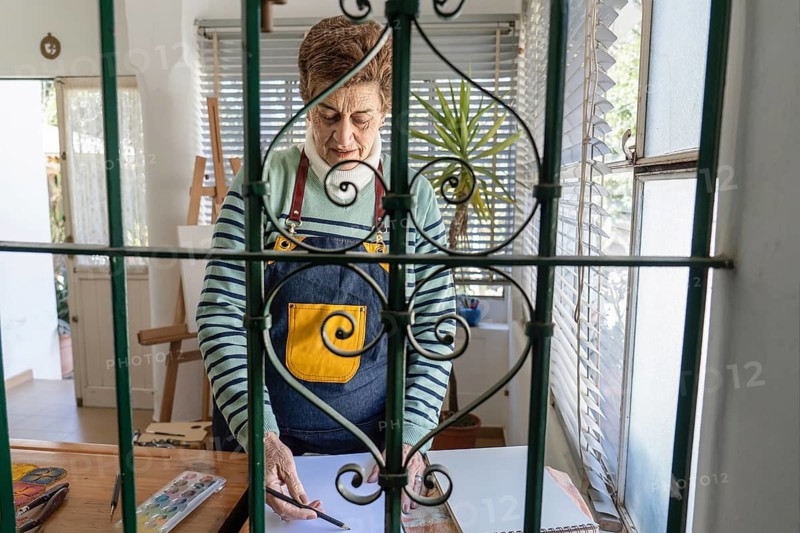 Creative framing of a mature female artist sketching at her art table, photographed through an ornamental green iron window grill, with watercolors and brushes visible on the table