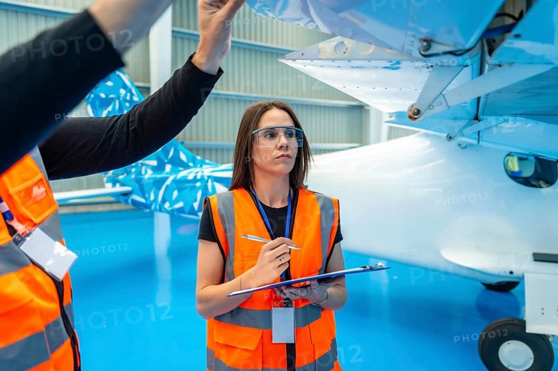 Female aviation engineer in safety vest and protective eyewear inspects an airplane wing section with clipboard and pen while a colleague points out a detail in a clean hangar