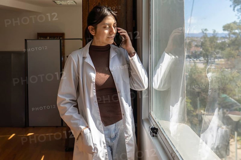 Young female scientist in white lab coat standing near a bright window while talking on a smartphone, looking outside with a calm expression and natural light on her face