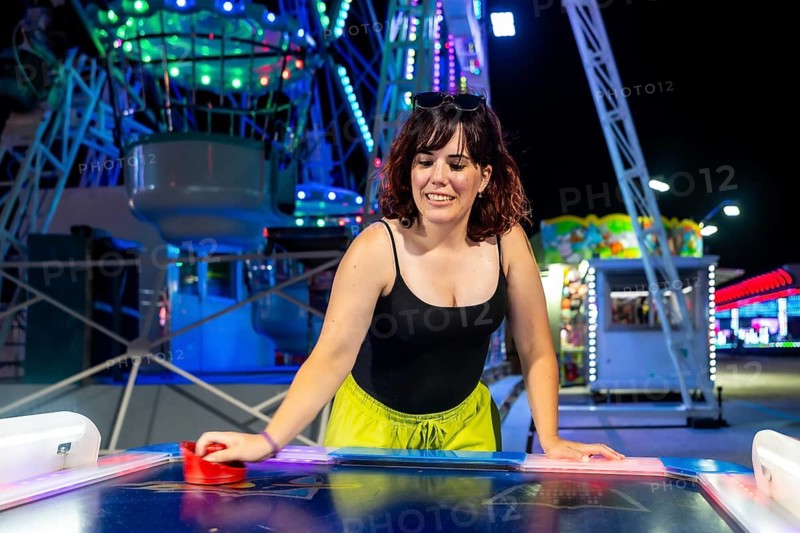 Young woman playing air hockey game at a vibrant night fair, smiling and concentrating on the competitive arcade activity illuminated by colorful lights of a ferris wheel