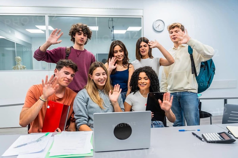 Group of young diverse students in a modern classroom actively participating in an online class or video call. Waving to the laptop screen. Showcasing teamwork. Technology. And contemporary education