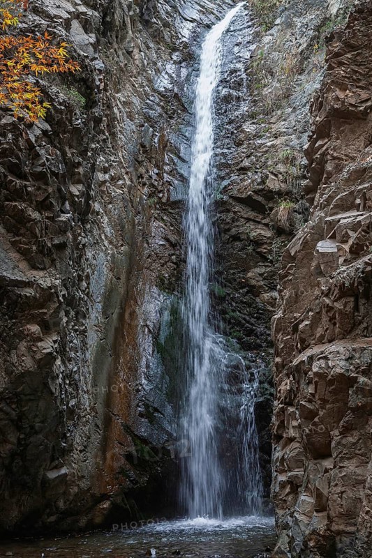 A high waterfall flows through a rocky gorge surrounded by autumn foliage and rough rocks, Millomeris Waterfall, Cyprus