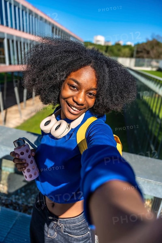 Smiling young student with afro hair, headphones, and a reusable water bottle, holding out a hand to take a selfie on an outdoor university campus during a sunny day