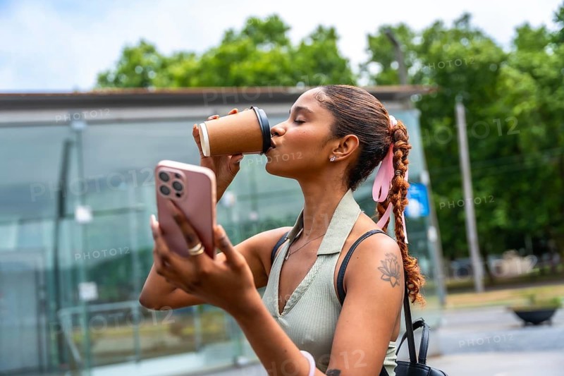 Young woman standing outdoors, wearing a casual outfit, enjoying a takeaway coffee while actively engaging with her personal smartphone, embodying modern urban lifestyle