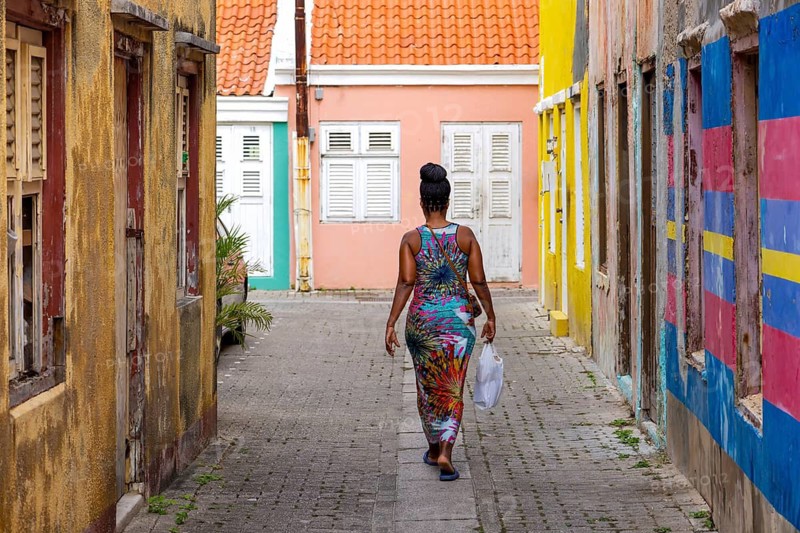 A rustic wooden pallet bench sits against a vibrant red colonial building wall with white shutters and pink flowers in Otrobanda district, Willemstad, Curacao