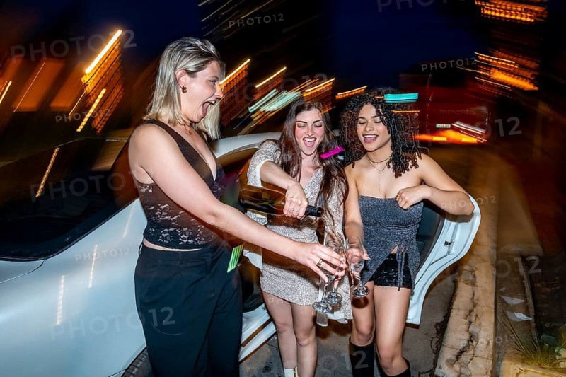 Three excited young women are celebrating a special occasion, pouring champagne into glasses at night outside a luxury limousine with city lights motion blurring in the background
