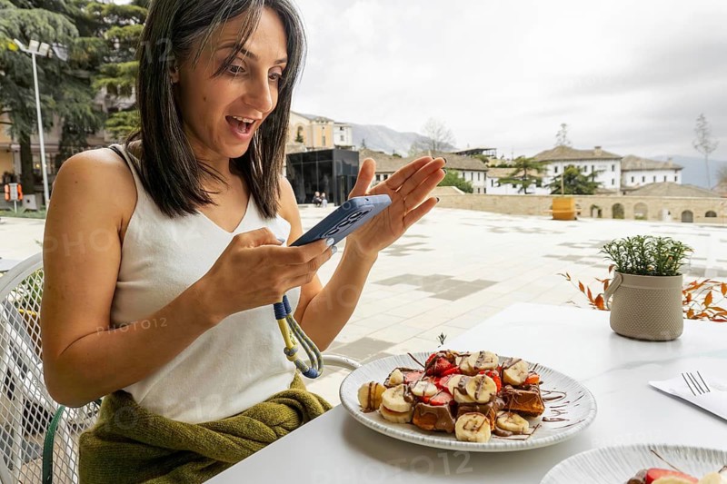 Excited slim woman traveler at an outdoor cafe in Gjirokastra hold use blue smartphone to photograph film capture delicious traditional waffle dessert served on table. Local fresh strawberry waffles