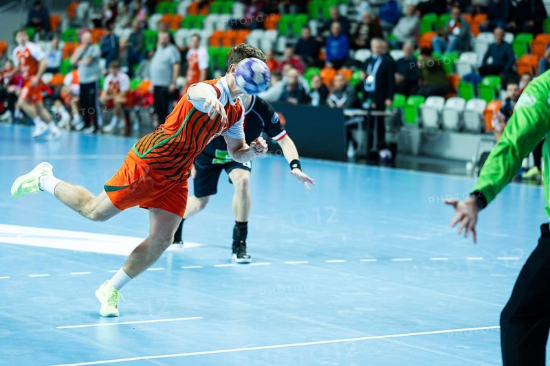 Professional handball player in orange jersey jumping and throwing ball during intense competitive match in indoor arena with blurred audience in background in Lubin, Poland