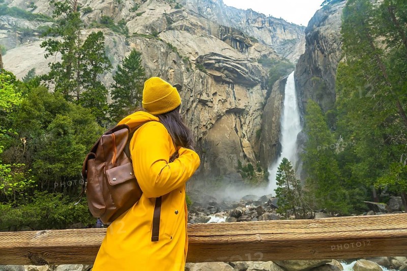 Woman wearing yellow rain jacket and beanie, carrying a backpack, standing on a wooden bridge enjoying the view of lower yosemite falls and rocky landscape in yosemite national park