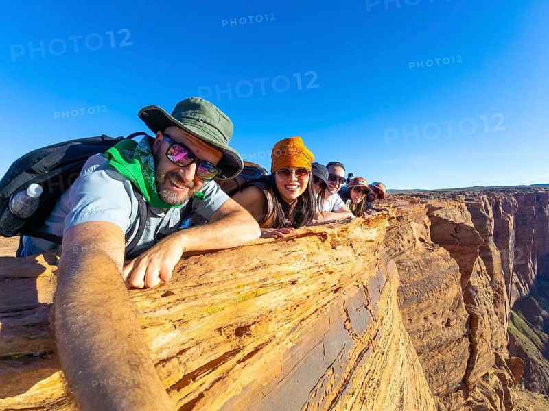 Group of diverse friends lying down on a cliff edge, smiling at the camera, enjoying travel and adventure at the iconic horseshoe bend in page, arizona