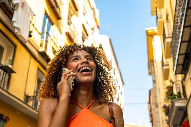 Happy young woman with curly brown hair laughing while talking on her mobile phone, enjoying a sunny urban street scene and a joyful, candid conversation during summer travel