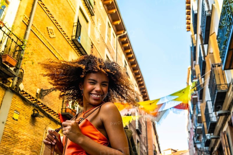Young woman with curly brown hair smiling at the camera on a vibrant madrid street, colorful festival banners overhead and historic buildings framing a sunny urban scene