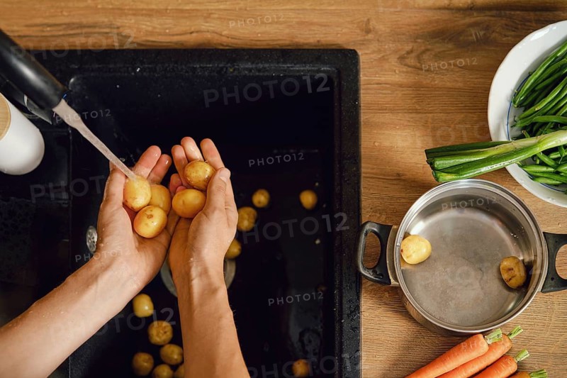 Hands hold baby potatoes under running water in kitchen sink. Nearby, there are fresh carrots and green vegetables on wooden surface. Pot waits for preparation beside sink