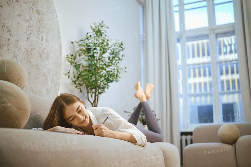 A woman lies on a sofa in a bright living room, using a pen to write notes. She has her feet up and is focused on her task. Sunlight streams through large windows illuminating the space