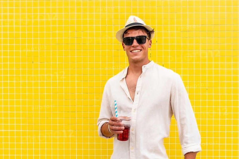 Smiling young man in a casual white linen shirt, fedora hat, and sunglasses holding a refreshing red drink with a straw, standing confidently against a vibrant yellow tiled background