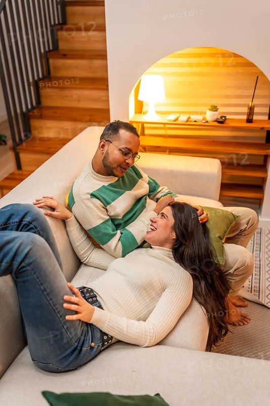 Happy couple relaxing together on a comfortable sofa in their modern living room, sharing a loving glance and smiling while enjoying a cozy evening at home