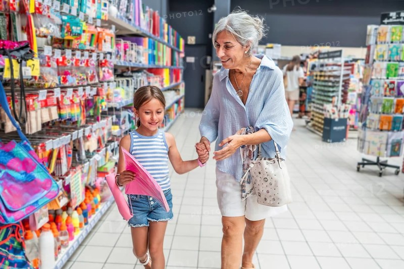 Grandmother and young granddaughter holding hands and smiling, walking down a vibrant aisle in a store, choosing colorful items for back to school while enjoying quality time