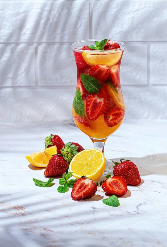 A tall glass filled with a colorful drink made from strawberries, lemon slices, and mint leaves. Fresh strawberries and lemon wedges surround the glass on a smooth surface