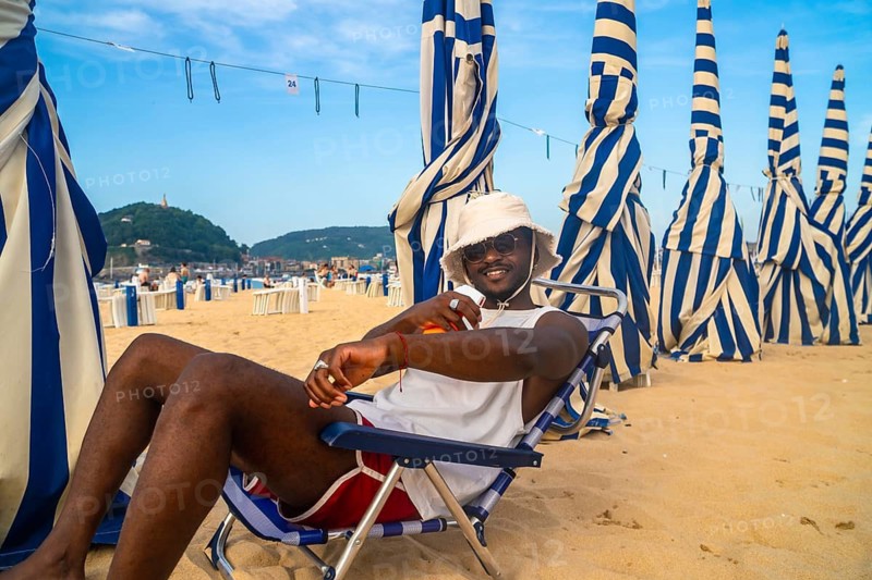 Young man wearing a bucket hat and sunglasses, sitting on a deck chair on a sandy beach under blue and white striped umbrellas, enjoying a sunny summer day