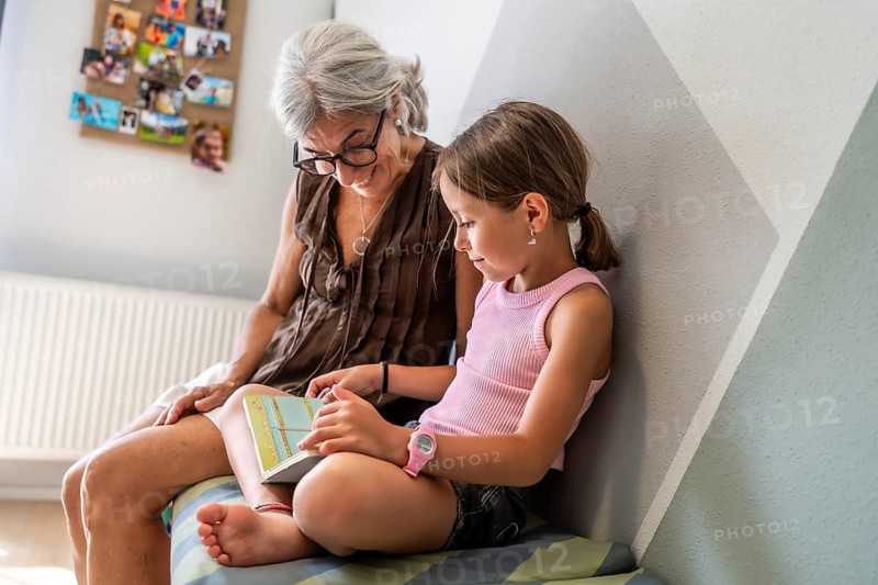Grandmother and granddaughter sitting together on a bench, looking at a children's book, sharing a moment of education and bonding at home