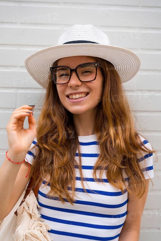 Young woman smiling broadly, wearing a white straw hat, black framed glasses, and a blue and white striped shirt, standing casually against a white brick wall