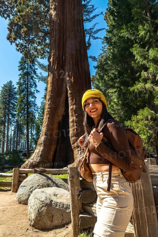 Happy young woman wearing a yellow beanie and backpack, smiling while exploring the majestic giant sequoia trees in a northern california national park on a sunny day