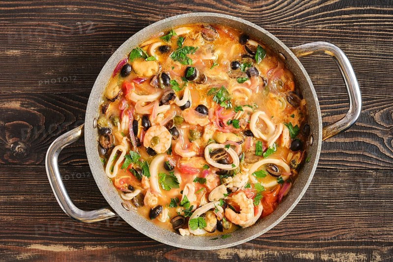 A pan contains a seafood dish with shrimp, squid, mussels and various vegetables. The meal is freshly cooked and shows a mix of colors. It is placed on a wooden table