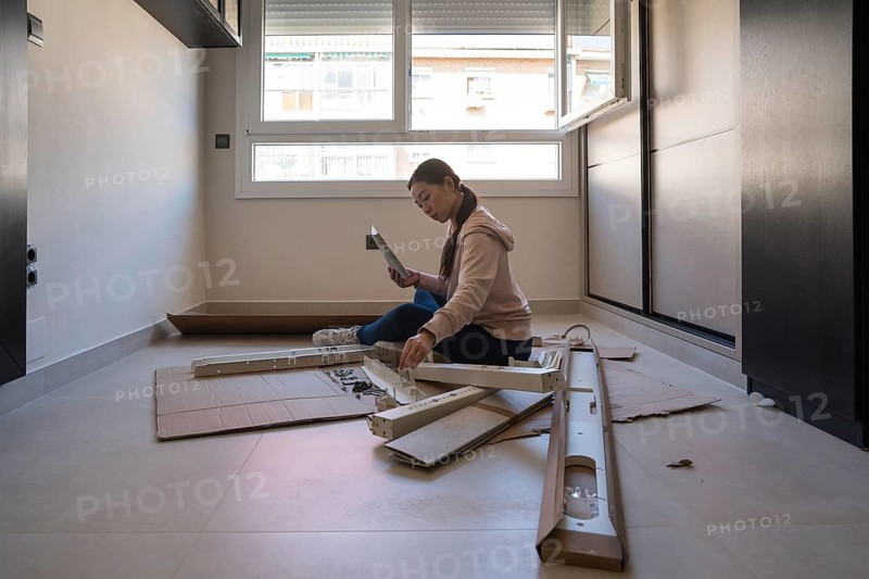 An Asian woman is focused on assembling a white desk at home, engaged in a DIY furniture project. The room is bright with sunlight illuminating her workspace