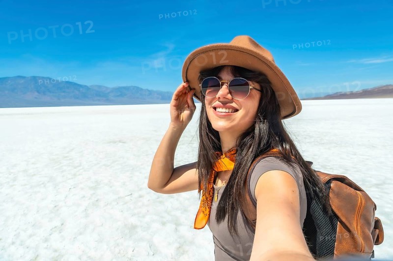 Happy young woman tourist enjoying a desert journey, smiling while taking a selfie at badwater basin, surrounded by the vast white salt flats and distant mountains under a clear blue sky
