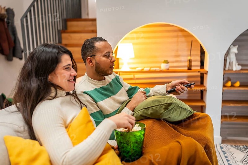 Happy young couple relaxing on a cozy sofa in their apartment, sharing popcorn and watching tv together on a weekend evening, wrapped in blankets and smiling