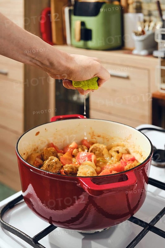 Hand is squeezing lime juice into pot filled with meat and vegetables on stove. Adding fresh sourness to food