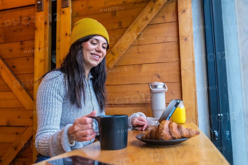 Woman in a cozy cafe enjoying breakfast with a croissant and coffee while browsing a tablet by the window, representing remote work and a digital nomad lifestyle
