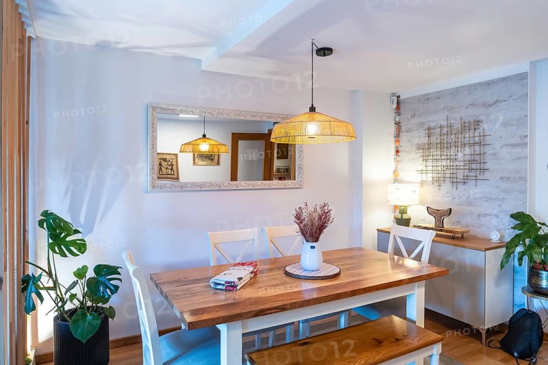 Modern dining room interior presenting a wooden table with white chairs and a bench, illuminated by a woven light fixture, contrasting with a mirrored wall and decorative plant
