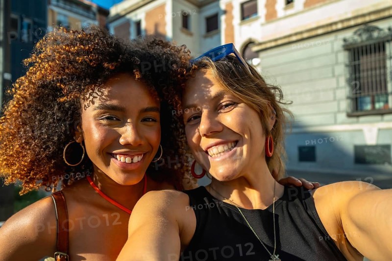 Two happy and diverse young women smiling at the camera for a selfie, celebrating friendship and connection during a sunny golden hour moment in an urban setting
