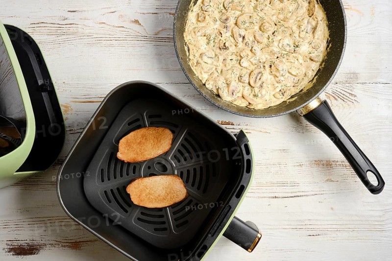 Two slices of meat are placed in air fryer basket. Skillet on tabletop contains creamy mushrooms. Cooking occurs in bright kitchen