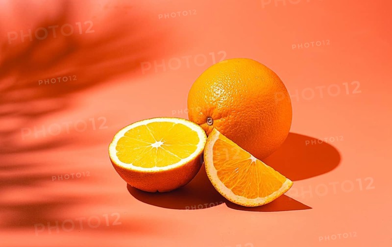 On the coral surface, next to two neatly sliced slices, there is a whole orange. Sunlight casts soft shadows, emphasizing the freshness and bright color of the fruit