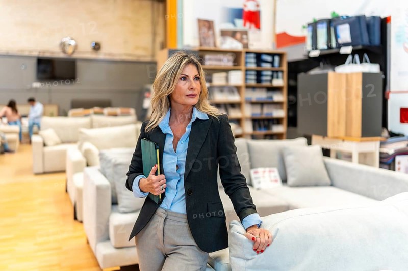 Businesswoman creating an inviting atmosphere while standing among modern sofas with a tablet, ready to assist customers browsing furniture in a home improvement store