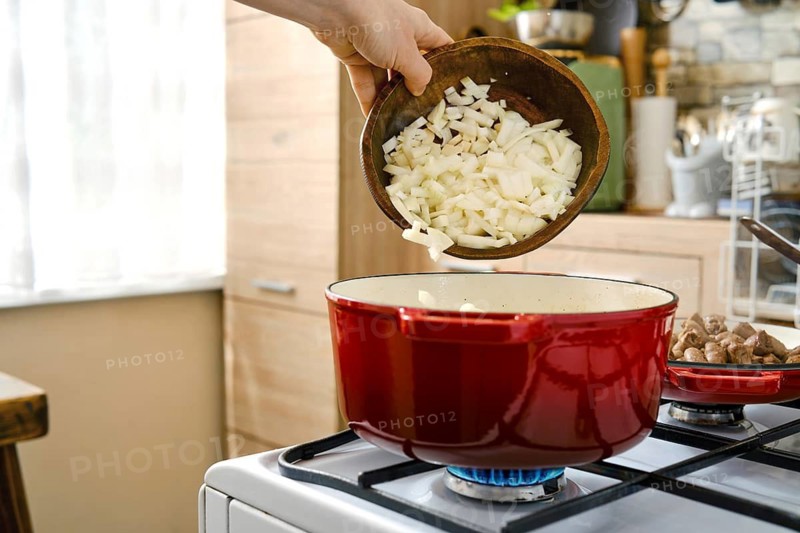 A hand transfers chopped onions from a bowl into a red pot placed on a stove. The kitchen has wooden cabinets and sunlight coming in through a window. It is daytime and food is being prepared