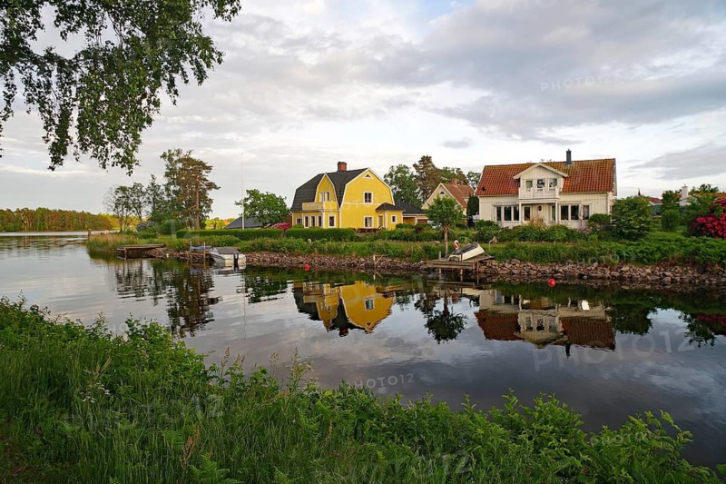 Two colorful houses by the river with a clear reflection in the water, surrounded by green nature and slightly cloudy sky, Karlsborg, Västra Götalands län, Sweden