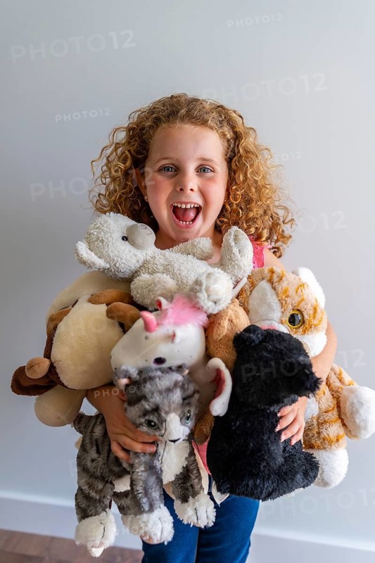 Little girl with curly hair laughing joyfully, holding a large pile of her beloved stuffed animals, embodying childhood happiness and the excitement of moving to a new home