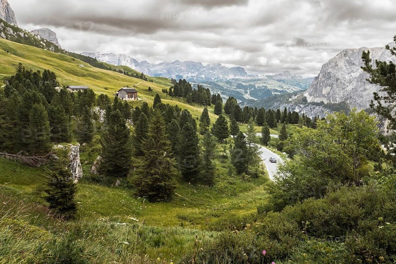 Aerial cinematic view car vehicle drives Winding mountain road through Dolomites landscape with alpine forest, rustic cabins, green meadows and dramatic peaks, ideal road trip travel concept