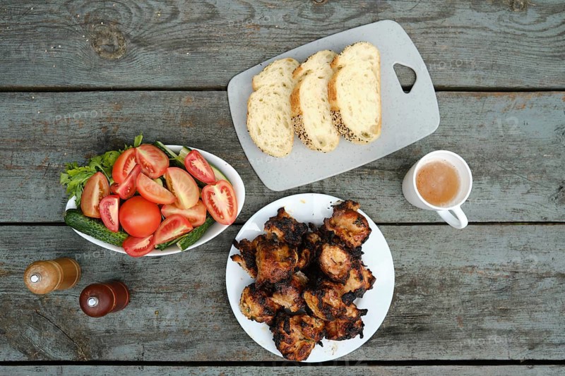 Grilled chicken placed on white plate alongside bowl of salad with tomatoes and cucumbers. Slices of bread sit on cutting board. Cup of juice is nearby
