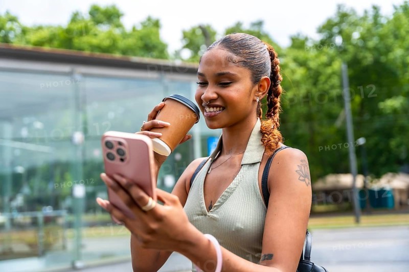 Happy young woman smiling while holding a reusable coffee cup and interacting with her cell phone in an urban outdoor setting, staying connected and enjoying a lifestyle moment