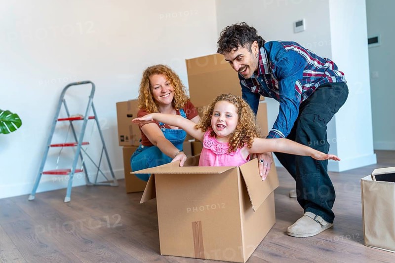 Happy family moving into a new home, parents pushing their smiling daughter inside a cardboard box, symbolizing fun, childhood joy, and the excitement of relocation for everyone