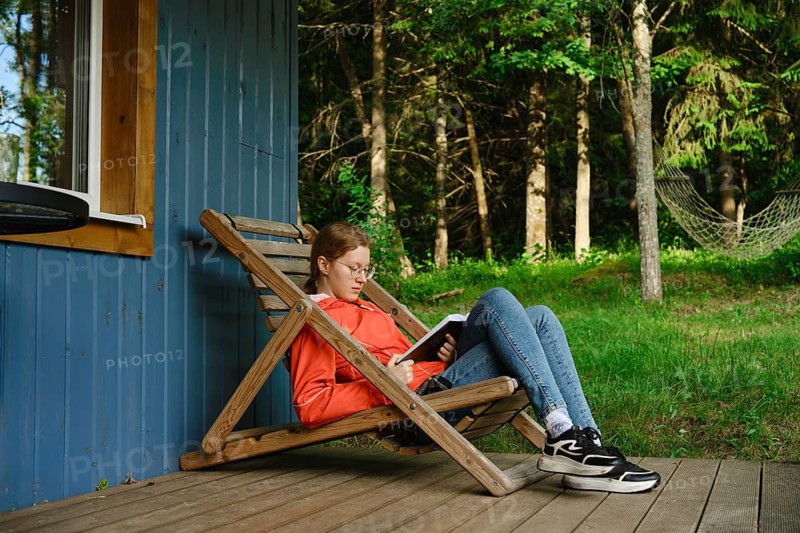 Girl sits on wooden chair on porch of wooden cabin reading book. Surrounding trees provide shade. Grass is green and lush. Sunlight filters through leaves. Calm moment during warm afternoon