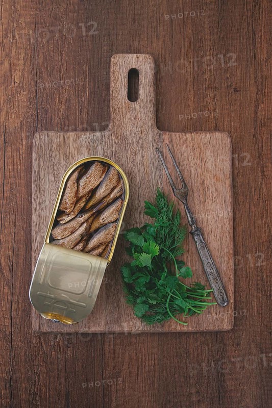 An open can of sprats, appetizer, on a wooden cutting board, with a fork and herbs, top view